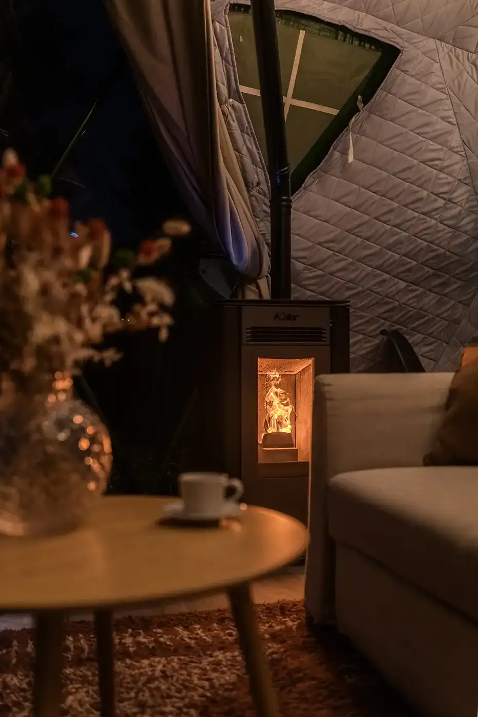 The lit pellet stove glowing in the evening, beside a sofa and a round table with a coffee and dried flowers