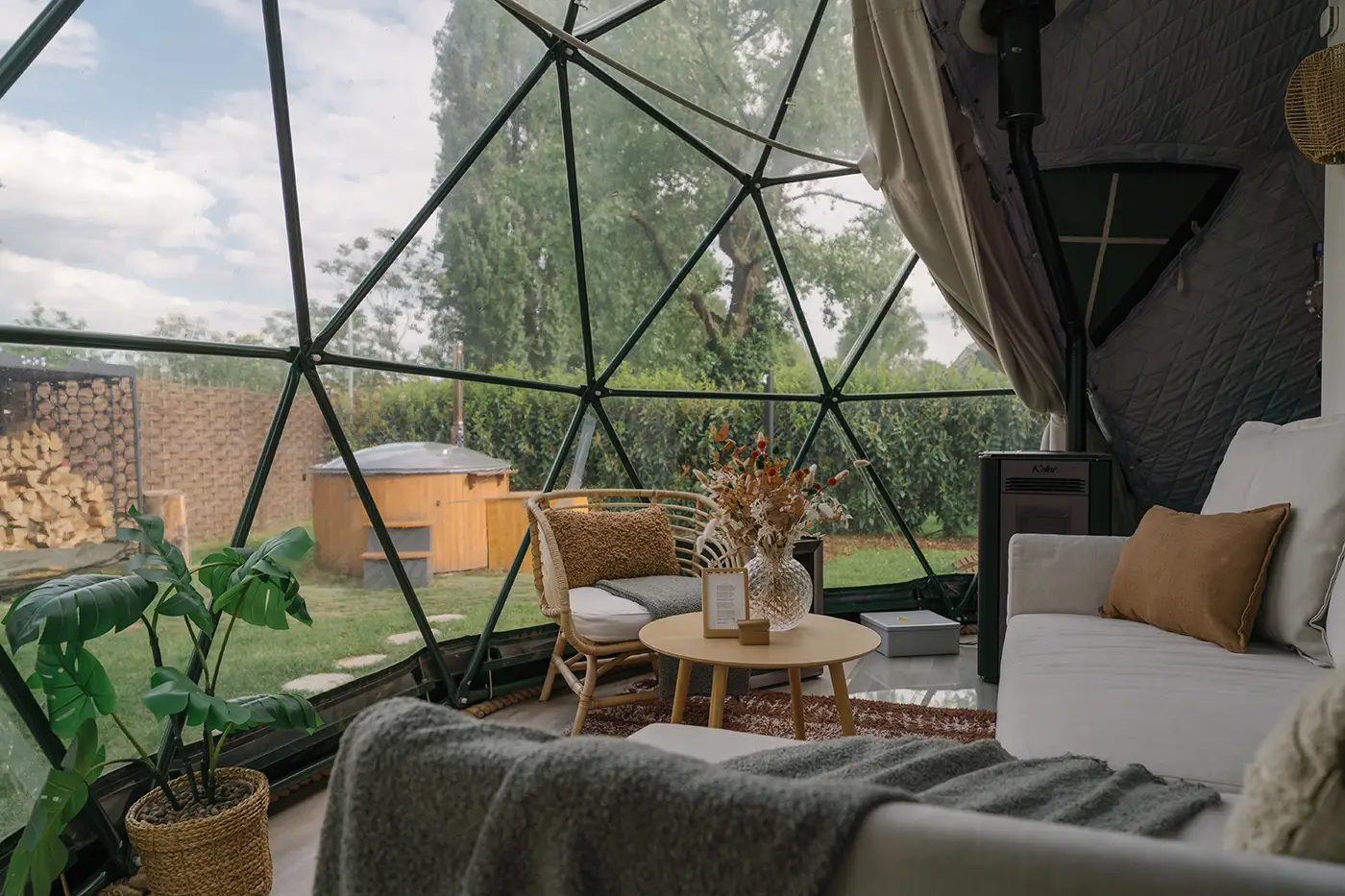 Inside the dome looking out through the glass facade to the garden, wood-fired hot tub and birch trees, with a sofa, armchair and pellet stove